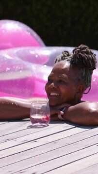 Vertical video: Leaning woman at pool edge, spotting person right, smiling with glass, straps