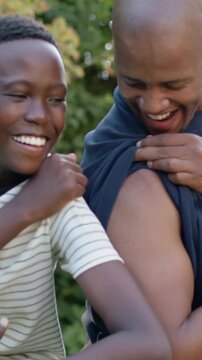 Vertical video: Lifting dark shirt, dad teasing son in striped tee, son touching bicep in garden