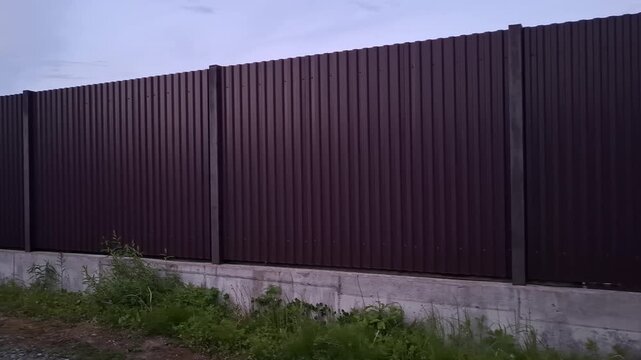 Brown corrugated metal sheet fence on concrete foundation. Modern iron boundary wall enclosing private property. Security, construction concept. Industrial profiled metal barrier on solid cement base