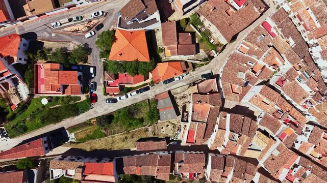 The scene shows a small town with houses having tiled roofs and narrow roads. Cars are parked along some streets. The shot captures the layout of the area from above.