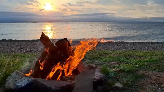 Campfire burning on pebble beach at sunset. Bright orange flames, calm sea, sun reflection on water, cloudy twilight sky. Beautiful bonfire blazing. Cozy relaxing atmosphere. Scenery lakeside campsite