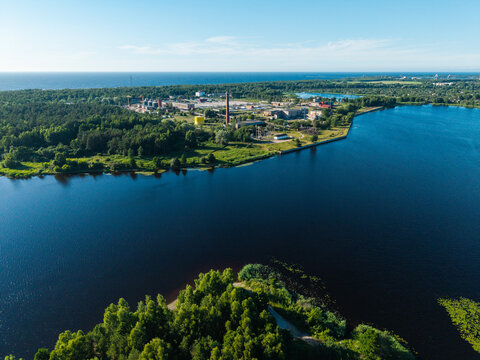 Aerial view of Daugavgriva featuring the confluence of the river and the sea, surrounded by dense forests, industrial buildings, and a tall chimney in Riga, Latvia.