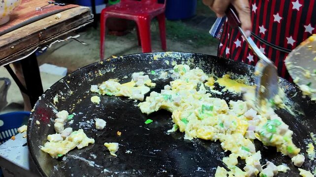 Thai street food vendor stir fries eggs with diced taro and chopped green onions on a hot flat griddle while preparing a traditional street food dish at a local night market in Thailand. Authentic Asi