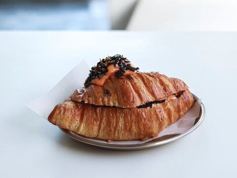 A savory croissant sandwich featuring a rich mentai sauce topped with shredded seaweed (nori) and sesame seeds, served on a plate against a soft, bright background.