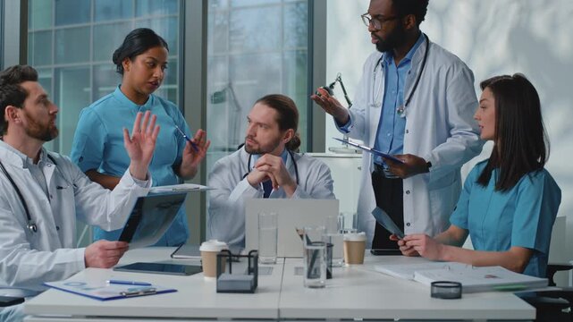 Stressed international five doctors colleagues arguing talking misunderstanding discussing health medical test bad results x-ray at table in hospital. Medicine team argue talk about trouble in clinic