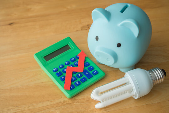 Piggy bank, light bulb, calculator with growth red graph on wooden table. Electricity bill prices increase, energy crisis in war, home financial management and planning, expense control concept.