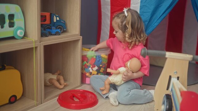 Girl holding a doll and reaching into a bin of colorful blocks inside a playroom building; playful curiosity.