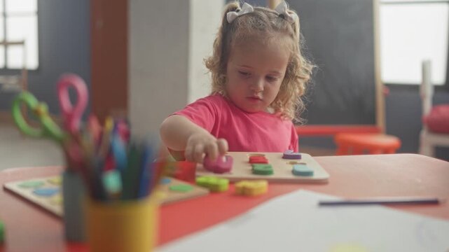 Girl places a yellow puzzle piece with her right hand on a wooden board at a classroom table, focused on shapes and colors; concentration learning curiosity.