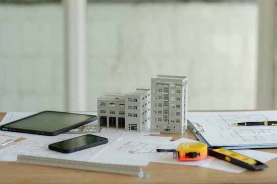 Close-up of architectural building models with blueprints, tablet, and construction tools on office desk.
