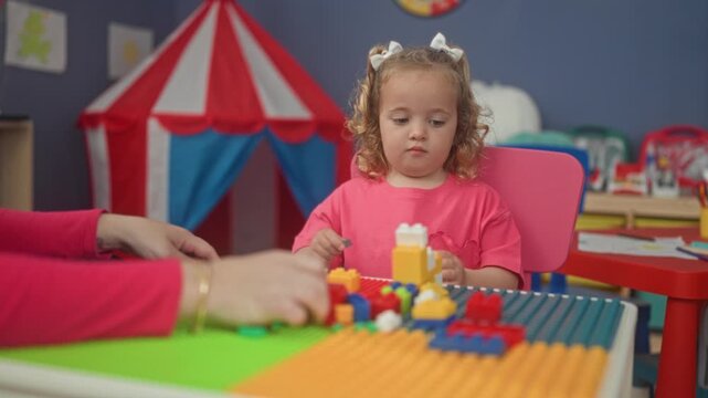 Girl reaches for colorful blocks on a play table inside a building playroom with an adult's hands nearby; childhood learning focus.