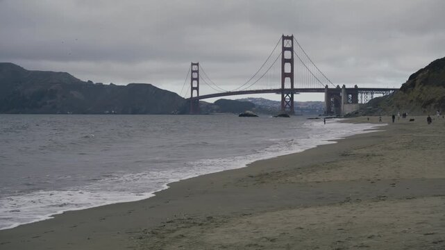 Wide Shot Of Golden Gate Bridge From Sandy Beach With People Walking On Overcast Day
