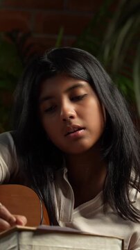 Indian Teenage Girl Playing Guitar While Sitting on a Bean Bag in Cozy Room with Moody Lamp Against Brick Wall, Composing or Writing Music as a Creative Hobby During Free Time at Home