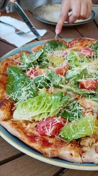Close up of freshly baked Caesar pizza with romaine lettuce and cherry tomatoes. Slicing Italian pizza on plate at cafe table.