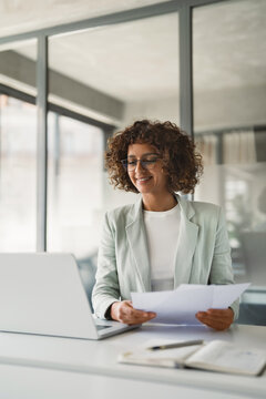 Latin hispanic young business woman working on laptop computer reading financial document report in office. Accountant entrepreneur manager businesswoman doing paperwork analysing using pc. Vertical