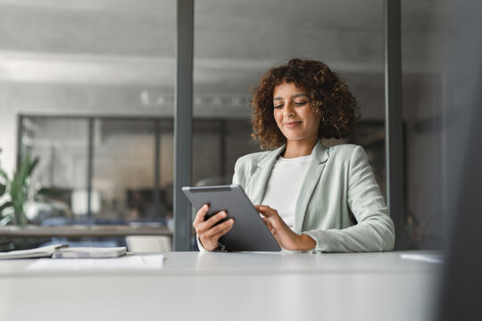 Smiling latin businesswoman sitting at office desk and using tablet at laptop in modern workspace, communication. Young indian woman holding tab screen device for work online. Technology and business