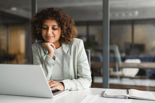 Young professional specialist latin hispanic businesswoman in suit working on laptop pc sitting at desk in office. Middle eastern indian woman using computer technology app for work online. Copy space
