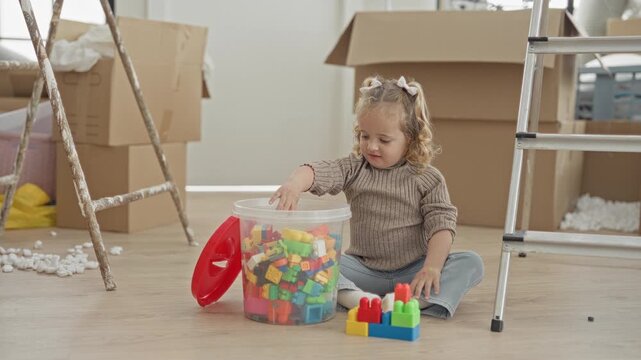 Little girl reaches into a clear plastic tub of colorful blocks with her hand, playing among moving boxes in a building; playful curiosity.