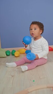 Little girl wearing white shirt and pink leggings puts a blue ball to her mouth while sitting on a wooden floor surrounded by colorful toy pins in a studio; curiosity discovery play.