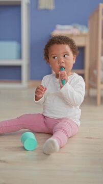 Little girl sitting on wooden floor near crib holding teal spoon with hand and touching mouth in building; curiosity innocence.