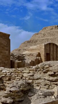 Step Pyramid of Saqqara, Egypt, designed by architect Imhotep for pharaoh Djoser, towering over archaeological ruins and stone debris under partly cloudy sky.