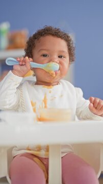 African american little girl eating with spoon and messy bib in highchair inside building; playful curiosity.