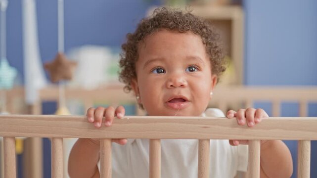 Little girl stands and grips crib rail with small hands in nursery building near mobile; tender curiosity warmth.
