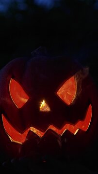 Vertical video. Glowing Halloween Pumpkin With a Scary Carved Face in Darkness Outdoors at Night. Holiday Spirit, Horror and Seasonal Celebration