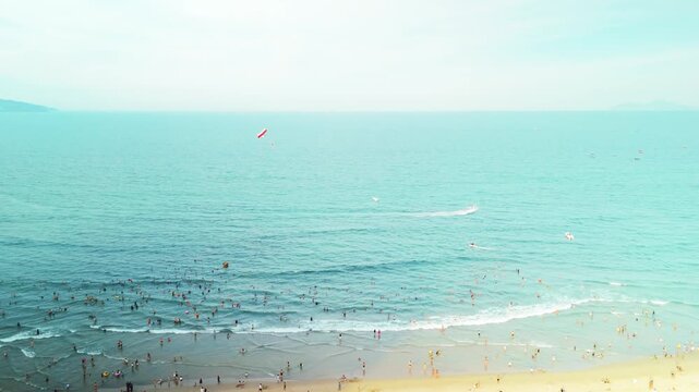 Da nang beach showing a crowded coastline with numerous people swimming and relaxing in the turquoise ocean water. While a parasailer glides above and speedboats create wakes