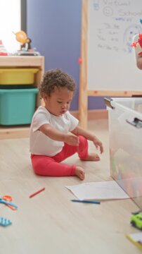 Little girl sitting on floor with red crayon and scattered paper, puts crayon to mouth near toy bin in playroom; curiosity.