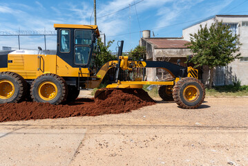 Fototapeta premium Side View of Motor Grader Machine Leveling Red Soil on Urban Street Construction Site