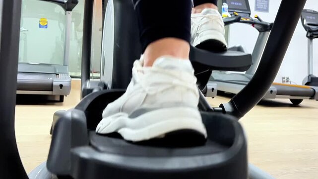 A person uses an elliptical trainer for exercise in a gym setting early in the day. The focus is on the feet in motion.