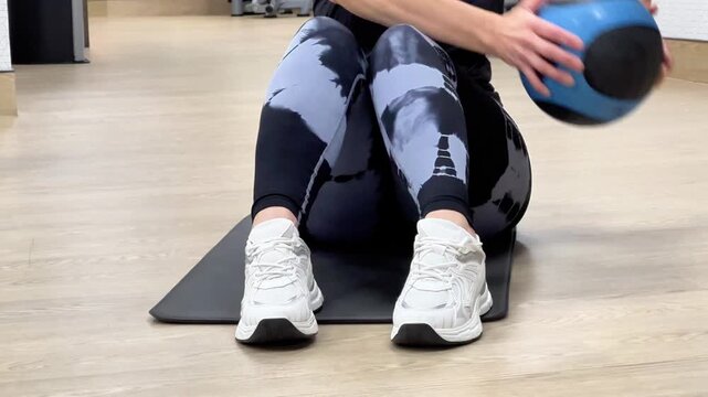 A person performs a medicine ball exercise on a mat in a gym. The activity focuses on core muscles and balance