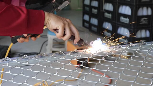 Welder wearing a protective face shield and gloves welds a steel rebar grid on the ground, with sparks and workshop tools visible in the background. Represents construction, metalwork, and industrial 