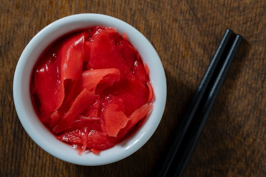 Pickled pink ginger in white bowl with black chopsticks. Top view marinated pink ginger slices in small white ceramic bowl next black chopsticks on rustic wooden table representing japanese cuisine