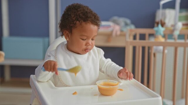 African american toddler girl feeding herself with spoon and bowl, tongue out in highchair in nursery building; playful curiosity.