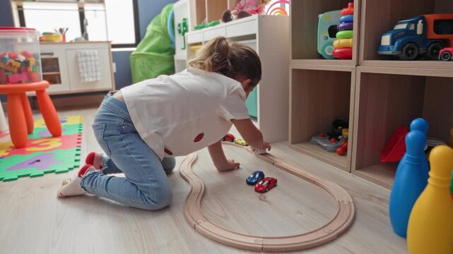 Hispanic girl kneeling on wooden floor, pushing a red toy car along a curved wooden train track beside cubby shelf, foam number mat and plastic pins in a playroom building; concentration.