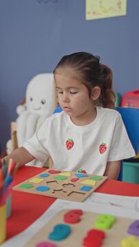 Girl placing wooden puzzle piece with hand at a red table in preschool classroom with toys and educational board; quiet concentration learning development.