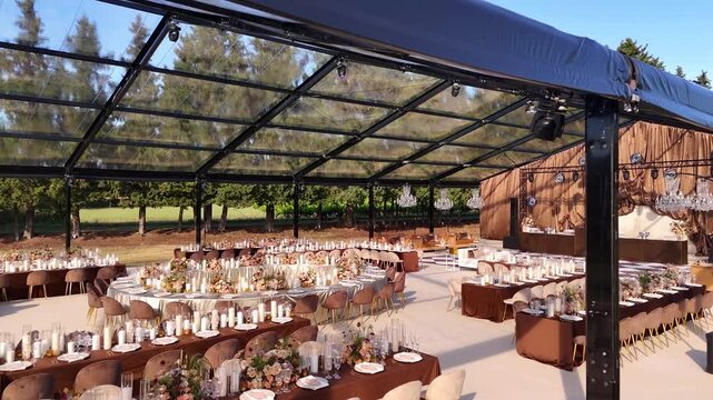 Luxury wedding reception setup with glass marquee and elegantly styled tables in milk beige and brown tones. Floral arrangements, candles, crystal chandeliers, and lighting for an evening celebration