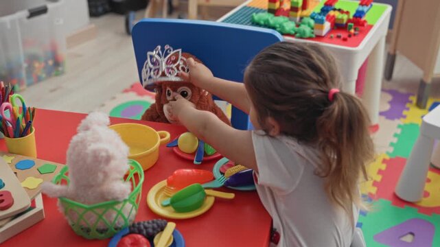 Hispanic girl placing tiara on stuffed monkey with outstretched arm at play table in building; imaginative play joy.
