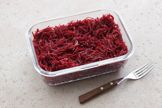 Fresh grated beetroot salad in a glass food container with fork on a light stone surface. Healthy vegetable dish, meal prep ingredient