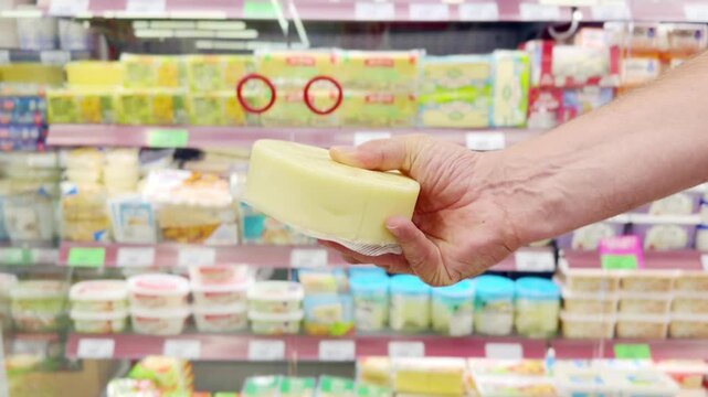 Customer's Hand Inspecting a Cheese Package While Shopping in a Grocery Store Aisle.Close-Up of a Man's Hand Spinning a Pack of Cheese While Browsing Products in a Supermarket Refrigerator Section