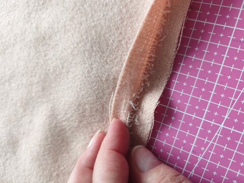 A top-down view of a seamstress or tailor hand-sewing and finishing the edge of a beige double-faced coat fabric.