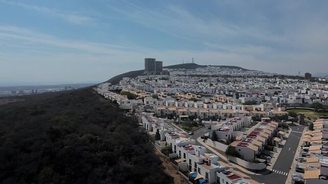 Drone forward approach of Zibata residential development displaying house neighborhoods apartment complexes hillside vegetation and Queretaro city backdrop