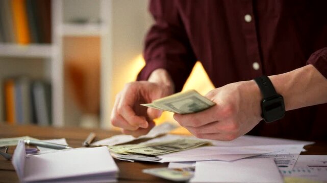 Man meticulously counts stacks of cash on cluttered table, immersed in work environment surrounded by scattered dollar bills, the atmosphere tense with financial focus as he prepares the day's budget