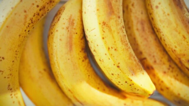 On sunlit kitchen table, bright banana branch rests, its yellow fruit speckled with brown spots, showing ripe summer produce. The close-up highlights natural ripening, evoking warm kitchen atmosphere