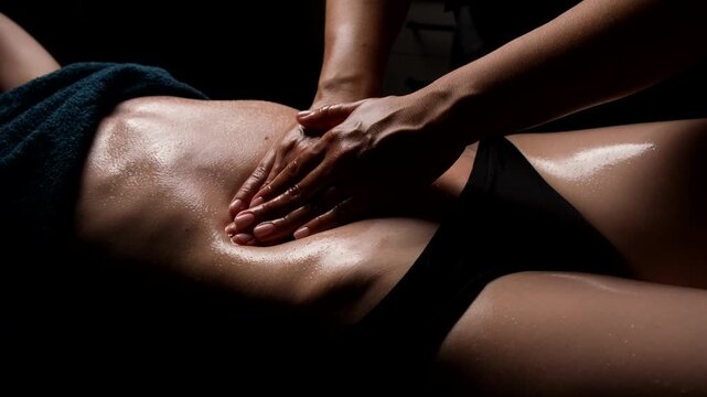 Skilled masseuse hands massaging a woman's abdomen with oil in a dark spa for lymphatic drainage