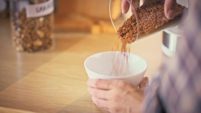 Pouring brown rice from a storage jar into a white bowl, a housewife works in her kitchen, preparing the grains for cooking. The simple ritual highlights daily meal prep in a homely setting