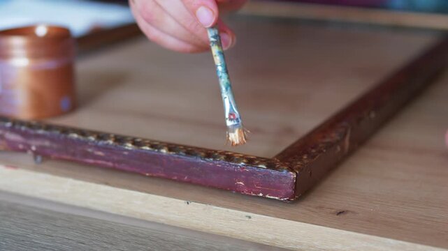 Close up of a hand applying metallic gold paint to restore an antique carved wooden picture frame