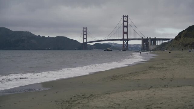 Golden Gate Bridge View From Sandy Beach As Ocean Waves Crash - Wide Shot on Stormy Day