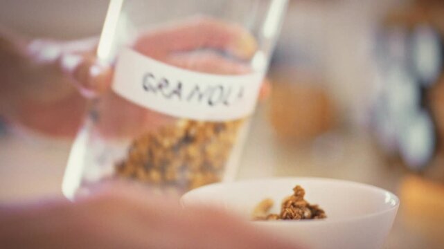 Woman's hands gently pour granola from storage jar into white bowl, crafting wholesome breakfast. The calm morning ritual highlights mindful nutrition, turning simple act into a balanced celebration